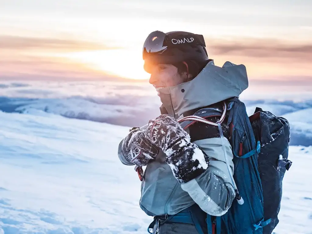 Une personne équipée pour le ski porte un casque, des gants et un sac à dos dans un paysage enneigé.