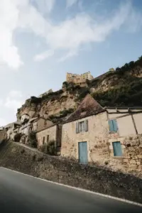 Maisons en pierre aux volets bleus, au pied d’une falaise surmontée d’un bâtiment fortifié.